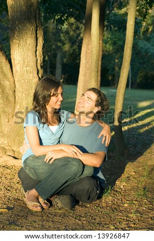 Happy, couple sit in the woods. She sits on his lap as he looks up at the sky. Vertically framed photograph