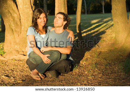 Happy,couple sit in the woods. She sits on his lap as he looks up at the sky. Horizontally framed photograph