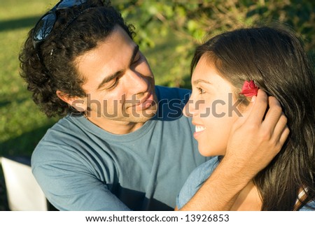 Happy couple on a park bench smile at each other as he places a red flower behind her ear. Horizontally framed photograph.