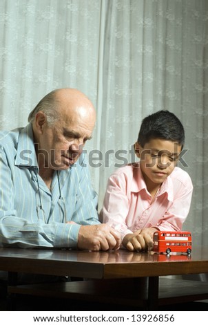 Grandfather and grandson are sitting at the table with a toy bus. Grandson pushes the toy bus softly as grandfather watches beside him. This is a vertically framed photo.