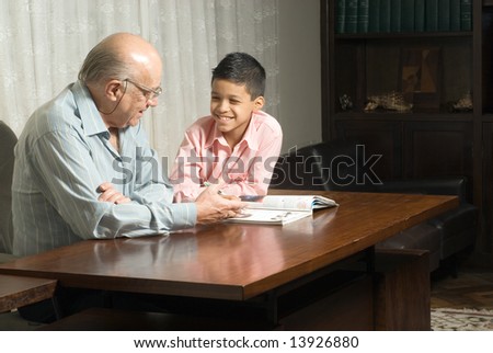 Grandfather and grandson are sitting at the table reading a book. Grandfather smiles as he reads the book to his grateful grandson. This is a horizontally framed photo.