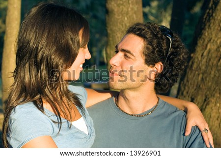 Couple staring at each other. She is smiling at him and he looks serious. Trees in the background. Horizontally framed photograph.