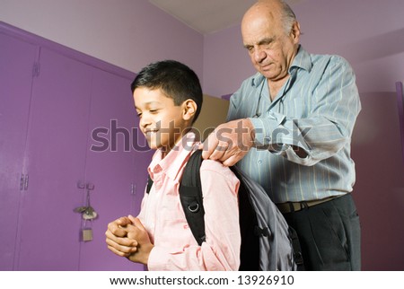 Grandfather is adjusting grandsons strap as he is leaving. Grandson stands smiling as grandfather adjusts his strap. This is a horizontally framed photo.