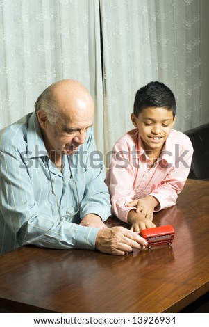 Grandfather and grandson sitting on the table playing with a toy bus. Grandfather shows the design on side of bus while grandson laughs. This is vertically framed.