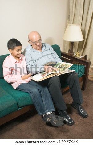 Grandfather and grandson sitting on couch together reading. Grandson reads book while grandson smiles and listens.  This is vertically framed.