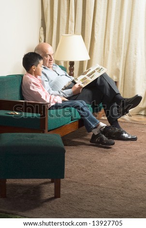 Grandfather and grandson sit on couch reading a book. Grandfather points to a picture on the page as grandson watches. This is a vertically framed photo.
