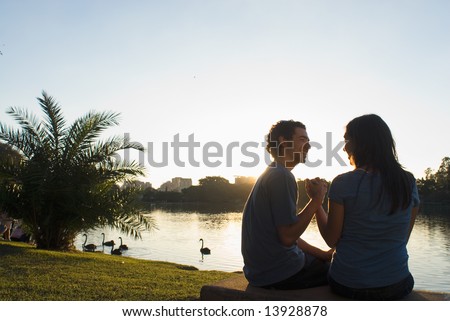 Happy couple clasp hands as they smile at each other. There is a pond with swans and buildings in the background. Horizontally framed photograph