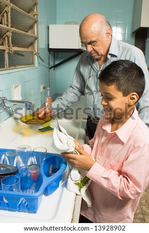 Grandfather and grandson are washing the dishes together. Grandfather washes a plate under the sink as grandson happily wipes down a utensil. This is a vertically framed photo.