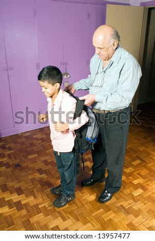 Grandfather is helping grandson put on his backpack as he gets ready for school. Grandson smiles while grandfather assists him with his backpack. This is a vertically framed photo.