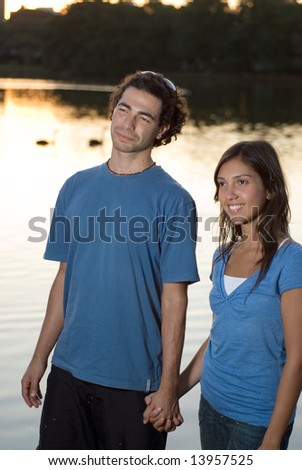 Couple holding hands and smiling. There is a pond and some trees behind them. Vertically framed photograph