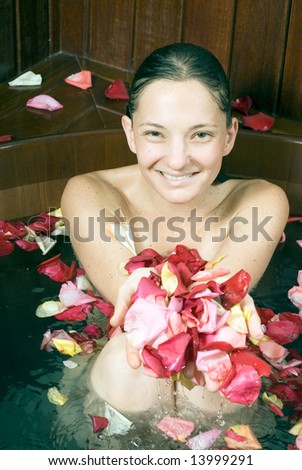 Attractive young women, covered in flower petals, soaking in a large dark-wood bathtub at spa and holding up her hands covered in petals.  Vertically framed close-up shot.