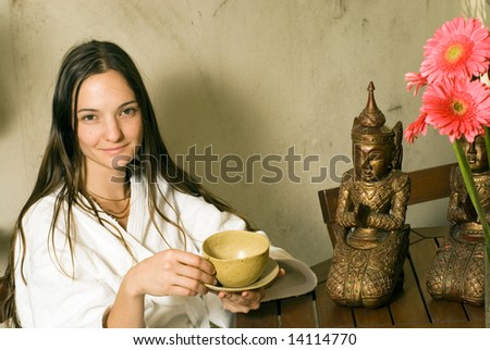 Woman smiles as she holds a tea cup. She sits next to a bronze statue and pink flowers. Horizontally framed photograph.