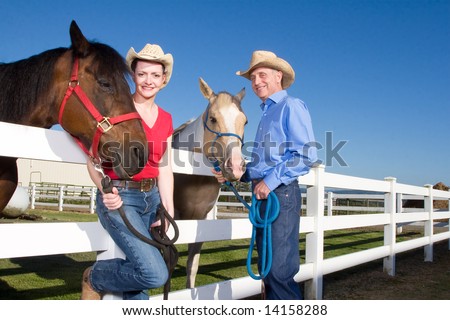 Smiling couple wearing cowboy hats stand next to their horses. She has her leg propped up on the fence. Horizontally framed photograph.