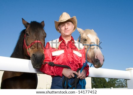 Cowboy in red shirt and a hat stands against a white fence in the middle of his two horses, one spotted blonde, the other brown. Horizontally framed photograph