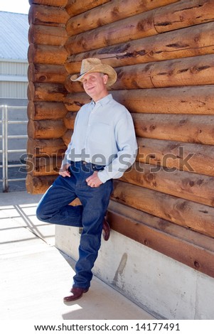 Man in cowboy hat leaning against a log cabin with his leg propped up. Vertically framed photograph