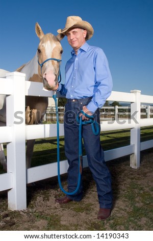 Cowboy smiles as he poses with his spotted horse next to a fence. Vertically framed photograph.