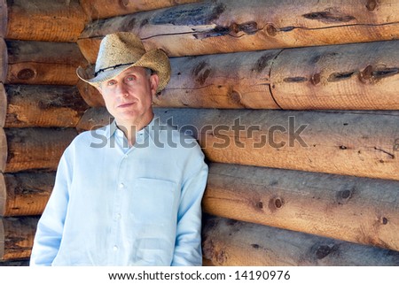Man in cowboy hat leaning against a log cabin. Horizontally framed photograph