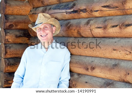 Laughing man in cowboy hat leaning against a log cabin. Horizontally framed photograph