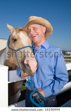 Cowboy laughs as he poses with his spotted horse next to a fence. Vertically framed photograph.