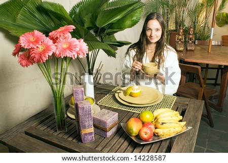 A young girl outside surrounded by plants, fruits, and flowers, holding a cup, smiling for the camera. - horizontally framed