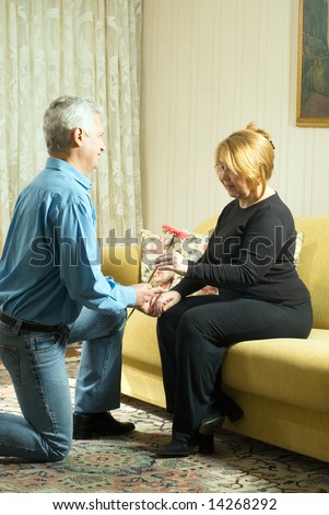 A man, on his knees, holding hands with his wife, sitting on the couch, offering a flower to her. - vertically framed