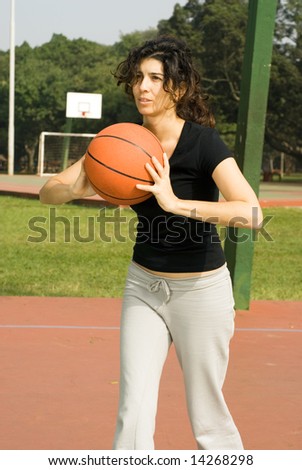 Young, attractive woman is standing on an outdoor basketball court.  She is holding a basketball and looking as if she is about to pass it to someone else.  Vertically framed shot.