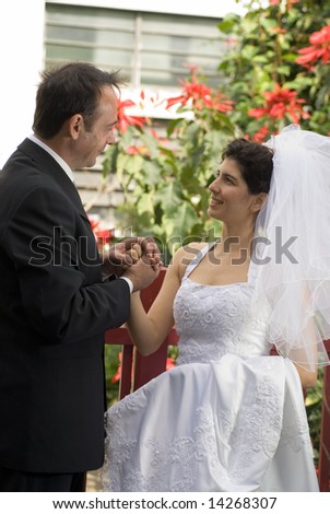 A newly married couple smile at each other while clasping hands, red flowers urban building with red flowers  in background. - vertically framed