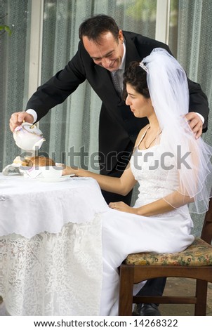 A newly married couple, the husband pours tea for the wife, while she sits on a white table-clothed table. - vertically framed