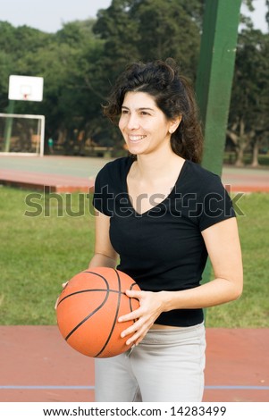 Young, attractive, happy woman is standing on an outdoor basketball court.  She is holding a basketball and looking in the other direction.  Vertically framed shot.