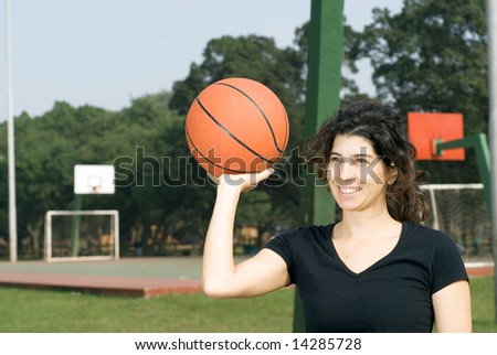Young, attractive woman is standing on an outdoor basketball court.  She is smiling and holding a basketball up with one hand.  Horizontally framed shot.