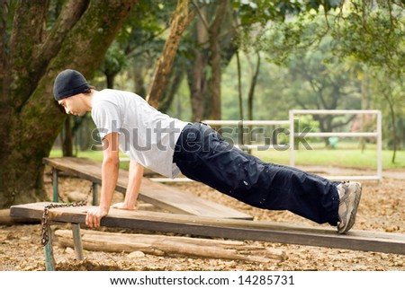 Man wearing hat is doing push-ups on top of a bench at a park. Horizontally framed shot.