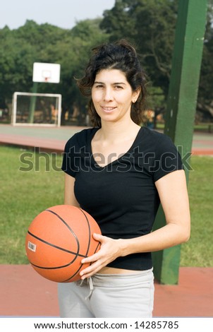Young, attractive, happy woman is standing on an outdoor basketball court.  She is holding a basketball and looking at the camera.  Vertically framed shot.
