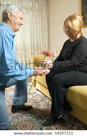 A man, on his knees, offering a pink flower, to a woman, on the couch. - vertically framed