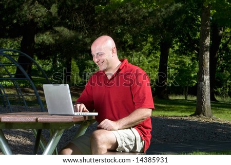 Bald gentleman seated on picnic table at the park smiling and playing on laptop computer. Horizontally framed shot.