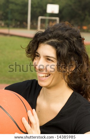 Young, attractive, happy woman is standing on an outdoor basketball court.  She is holding a basketball and smiling and laughing.  Vertically framed shot.
