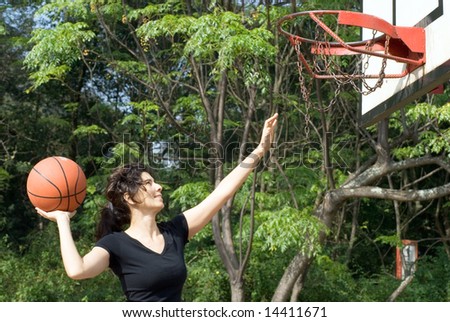 A woman is playing basketball on a court at the park.  The woman is looking at the basketball hoop and about to make a shot.  Horizontally framed photo.