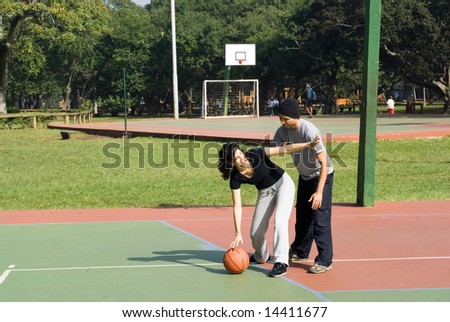 A man and a woman are playing on a park basketball court.  The woman is dribbling the basketball and blocking the man.  They are looking away from the camera.  Horizontally framed shot.