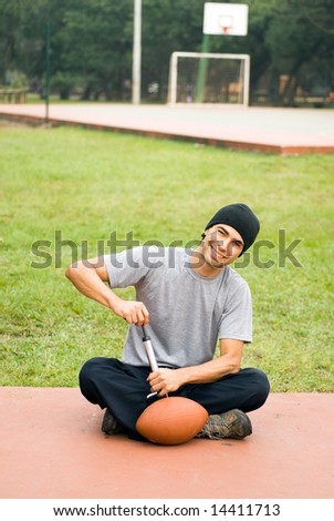 A man is sitting in a park.  He is smiling, looking at the camera and pumping air into a football.  Vertically framed photo.