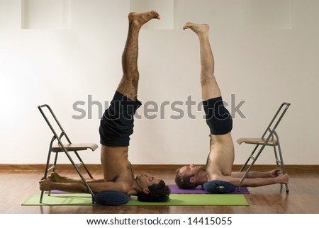 Two men exercising. Lying on their backs with their legs pointed upwards, and their arms holding chairs. Horizontally framed shot.