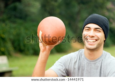 A man, sitting on the park ground, holds a football with one hand, smiling - horizontally framed
