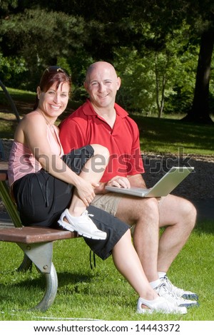 Couple lovingly sitting on a picnic table smiling and sitting at the park. The man is holding a laptop computer. Vertically framed shot.