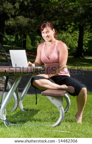 Woman smiles as she works on her laptop on a park bench. Vertically framed photograph