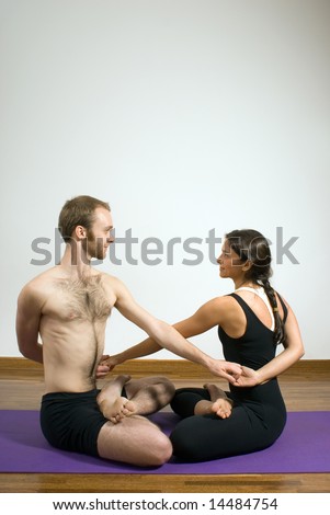 Man and woman sit on the floor on yoga mats in a yoga position with their arms entwined and their legs crossed. Vertically framed photograph