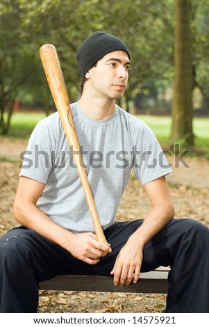 A man, sitting on a park bench, poses with a wooden baseball bat - vertically framed