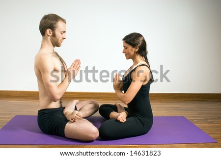 Man and woman performing yoga together on one large mat. Facing each other, eyes closed, legs crossed, and hands in prayer formation. Horizontally framed shot.