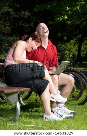 Couple lovingly sitting on a park bench laughing together. The man is holding a laptop computer and the woman is resting on his shoulder. Vertically framed shot.
