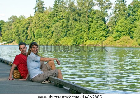 A couple, sit together on a dock, with their feet dangling over, smiling. - horizontally framed