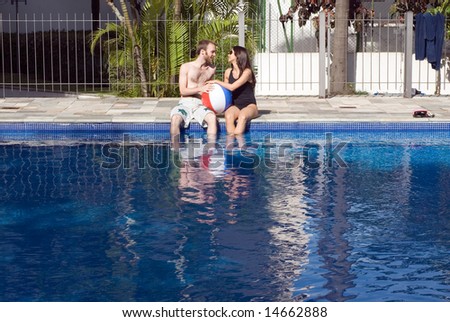 A man and a woman are relaxing together beside a pool.  They are looking at each other and dipping their legs in the water.  They are holding a beach-ball, Horizontally framed photo.