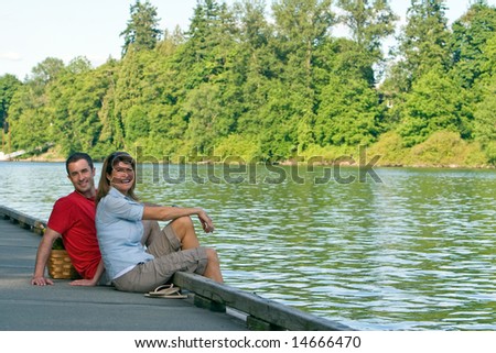 A couple, sit together on a dock, with their feet dangling over, smiling. - horizontally framed