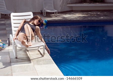 A woman, lying in a pool chair, smiling and relaxing, dips her hand into the pool. - horizontally framed